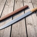 Hand forged 1095 Viking seax with a long single edged blade and Elder Futhark runes lying on a weathered picnic table beside its tan leather sheath