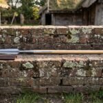 Hand forged 1095 Viking spear with a leaf shaped head and rune engraved socket resting on a lichen covered brick wall in front of a turf roofed longhouse