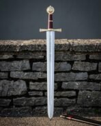 Hand forged arming sword with a brass wheel pommel and oxblood leather grip standing point down against a dry stone wall beside its red and black scabbard