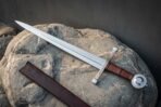 Hand forged arming sword with a polished fullered blade, knotwork engraved crossguard, leather wrapped grip and disc pommel, lying on a boulder beside its brown leather scabbard.