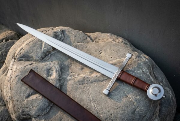 Hand forged arming sword with a polished fullered blade, knotwork engraved crossguard, leather wrapped grip and disc pommel, lying on a boulder beside its brown leather scabbard.