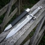 Hand forged bayonet with fullered blade and ribbed steel grip angled on an old rail next to its black scabbard