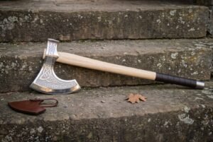 Hand forged bearded axe in bright 1095 steel with etched knotwork and rune band, pale ash haft and leather grip, lying on weathered stone steps beside its sheath