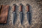 Hand forged black coated throwing knives with paracord grips, three standing point down in a row on gravel beside a leather roll