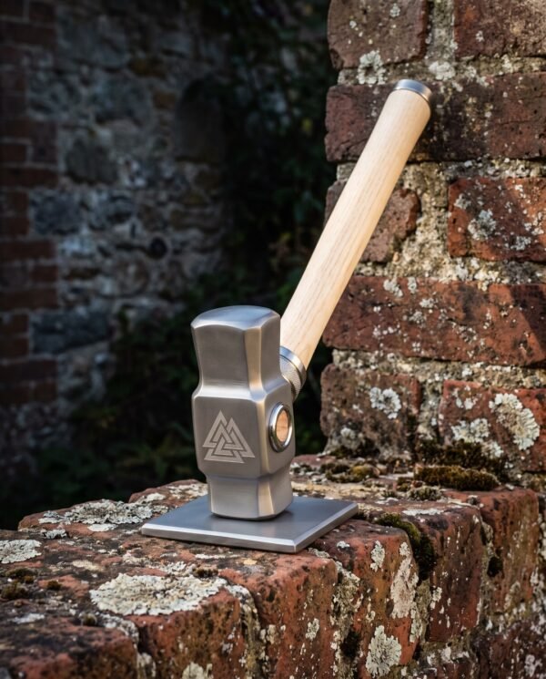 Hand forged blacksmith hammer with an octagonal 1095 carbon steel head, engraved Valknut on the cheek and an American ash handle, standing on a weathered brick wall