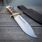 Hand forged Bowie knife with brass double guard and stacked leather handle on grey boards beside a brown leather sheath