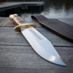 Hand forged Bowie knife with brass guard shown close by the water on wooden planks, wide clip point blade and sheath