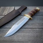 Bowie knife with etched blade, stacked leather grip and brass guard lying on decking with its brown belt sheath above