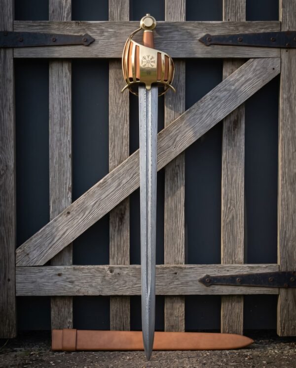 Hand forged brass basket hilt sword against an old wooden gate, damascus blade below a Vegvisir engraved brass cage, tan leather grip and matching leather scabbard