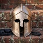 Hand forged bronze toned Greek helmet in the Corinthian style, hammered crown and engraved Greek key border, photographed on weathered red brick steps in daylight.