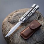 Hand forged butterfly knife with skeletonised steel handles fully open on a rounded rock beside its leather belt pouch