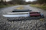 Hand forged chain sword pair with oxblood grips and ring pommels on blackened chains laid on a gravel track with scabbards