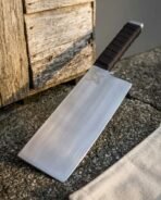 Hand forged cleaver with straight edge and cord wrapped handle angled on gravel against a wooden wall, canvas roll in view