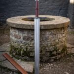 Hand forged damascus arming sword standing at an old stone well, layered blade below a blued crossguard, banded burgundy grip and a Celtic knot wheel pommel
