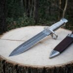 Hand forged Damascus bayonet dagger with fluted steel grip and ring guard lying diagonally on a log slice beside its sheath