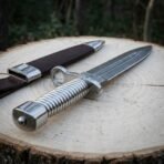Hand forged Damascus bayonet dagger with ring guard and fluted steel grip across a tree stump in front of its brown sheath