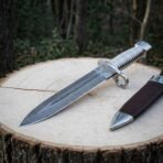 Hand forged damascus bayonet knife with a fluted polished steel grip, ring guard and triskele engraving lying on a pale wood stump beside its brown leather sheath