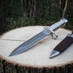 Hand forged damascus bayonet knife with a fluted polished steel grip, ring guard and triskele engraving lying on a pale wood stump beside its brown leather sheath