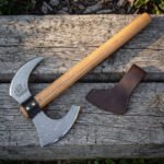 Hand forged Damascus bearded axe with engraved longship and hooked horn, full length on a wooden sleeper beside its blade mask
