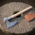 Hand forged Damascus bearded axe with pale ash haft and steel collar, close angled view on a carved millstone with leather cover