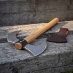 Hand forged Damascus bearded axe with riveted leather collar and hooked horn, close angled view of the head on timber