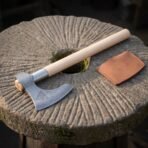 Hand forged Damascus bearded axe with triquetra engraved cheek and pale ash haft resting on a millstone beside its leather cover