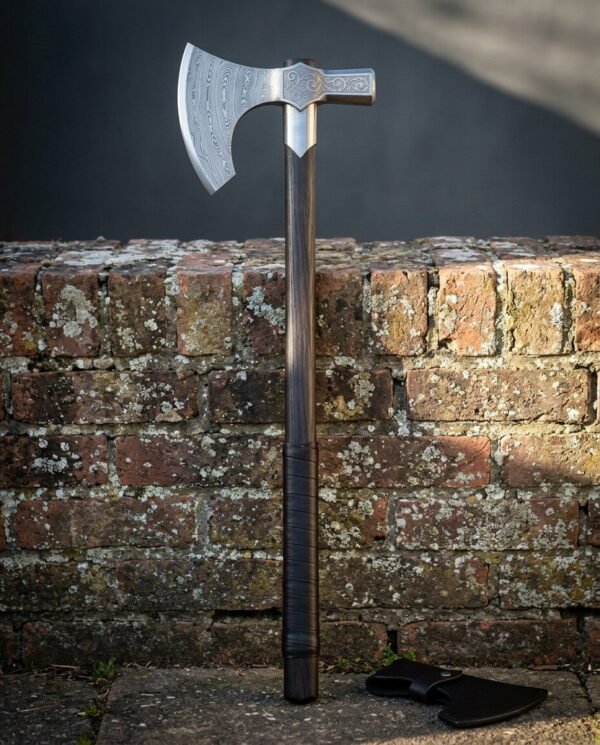 Hand forged damascus bearded axe with an engraved hammer poll and a leather wrapped dark hardwood haft standing against a lichen covered brick wall in evening light