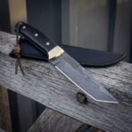 Hand forged Damascus blade with brass bolster and black scales pointing down on a fence rail, lanyard hanging from the handle