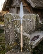 Hand forged damascus boar spear with a broad leaf blade, swept steel crossbar wings, brass knotwork ferrule and ash haft standing against a stone bench outside a timber longhouse