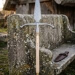 Hand forged damascus boar spear with a broad leaf blade, swept steel crossbar wings, brass knotwork ferrule and ash haft standing against a stone bench outside a timber longhouse