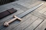 Hand forged Damascus bone handle Viking sword with brass lobed pommel and ribbed grip lying beside a brown leather scabbard on weathered grey decking boards