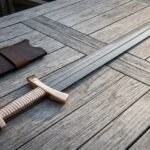 Hand forged Damascus bone handle Viking sword with brass lobed pommel and ribbed grip lying beside a brown leather scabbard on weathered grey decking boards