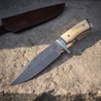 Hand forged Damascus bowie knife with bone handle scales, angled view from handle to tip with the oxblood sheath behind