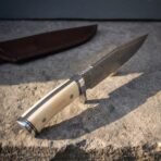 Hand forged Damascus bowie knife with bone handle scales, close view of the handle and blade spine in low sunlight on stone