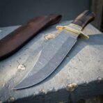 Hand forged Damascus bowie knife with ram horn handle and brass guard, close view of the blade tip under its leather sheath