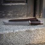 Hand forged Damascus bowie knife with stacked leather handle and clip point, full side view on a granite step by a wooden door