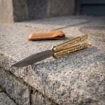 Hand forged Damascus butterfly knife with brass handles open near the edge of a granite step, tan leather pouch resting further back