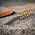 Hand forged Damascus butterfly knife with skeletonised brass handles, close angled view of the blade pattern with its tan leather pouch behind