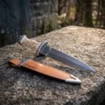 Hand forged Damascus Celtic dagger with carved bone handle, low angle view along the blade above a tan leather sheath on stone