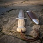 Hand forged Damascus Celtic dagger with carved bone handle seen from the pommel end, leather cord and brown sheath on stone