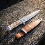 Hand forged Damascus Celtic dagger with carved bone handle and spiral etched ricasso pointing upward beside its tan leather sheath on granite