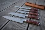 Hand forged Damascus chef knife set of five with rosewood handles stacked diagonally on grey decking below a leather knife roll