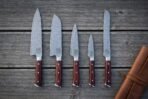Hand forged damascus chef knife set of five with rosewood handles and Vegvisir etched blades laid out on grey decking beside a rolled tan leather knife roll
