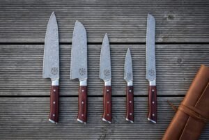 Hand forged damascus chef knife set of five with rosewood handles and Vegvisir etched blades laid out on grey decking beside a rolled tan leather knife roll