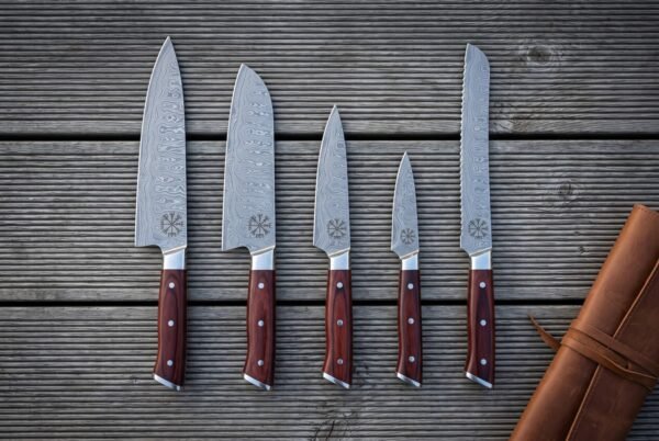 Hand forged damascus chef knife set of five with rosewood handles and Vegvisir etched blades laid out on grey decking beside a rolled tan leather knife roll