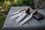 Hand forged Damascus chef knife set with walnut handles and brass bolsters fanned on stone with the leather roll, greenery behind