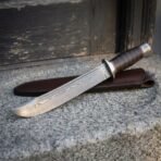 Hand forged Damascus clip point bowie with fullered blade, close angled view on granite with the dark leather sheath behind