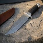 Hand forged Damascus combat knife with cord wrapped grip, close view of the patterned blade and guard on pitted stone