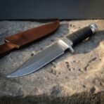 Hand forged Damascus combat knife with cord wrapped grip and steel guard, angled view along the blade with its leather sheath behind