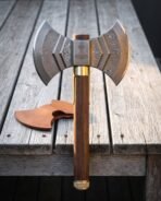 Hand forged Damascus double headed axe with layered twin crescent bits, an engraved Mjolnir cheek, walnut haft and brass collar standing on a weathered wooden deck